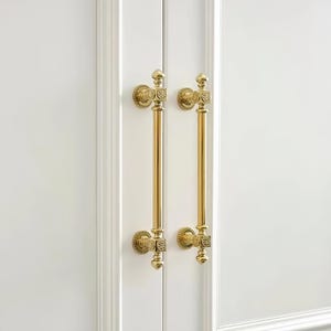 May include: Two vertical brass door handles with a polished gold finish, featuring square mounting blocks with circular floral engravings and decorative finials at the top and bottom, mounted on a white paneled door with vertical trim details.