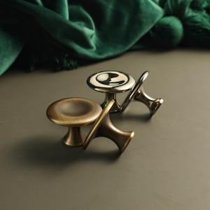 May include: Three metal cabinet knobs with a circular, concave top and a tapered base. One knob features a polished, reflective silver finish, while the other two knobs have a brushed, antique brass finish with visible metallic texture.