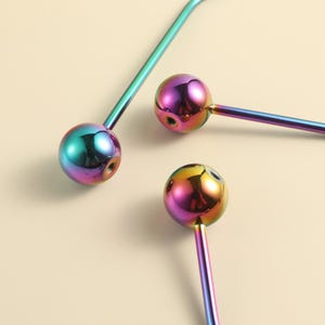 Puede incluir: Tres piezas de joyería corporal de metal con acabado iridiscente de arcoíris, cada una con un extremo esférico liso unido a un tallo de metal fino y curvado. El acabado metálico muestra un degradado de tonos azules, verdes, morados y dorados.