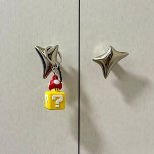 May include: A silver-colored star-shaped cabinet knob with a metal keyring attached. A small yellow cube-shaped block with a white question mark and a red and white mushroom figure are suspended from the keyring by a silver metal chain.
