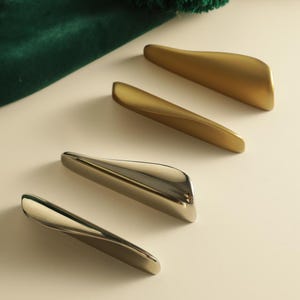Könnte beinhalten: Vier längliche Schrankgriffe, die in einer diagonalen Reihe angeordnet sind. Zwei Griffe haben ein mattes Gold-Finish, während die anderen beiden ein poliertes Silber-Finish aufweisen. Jeder Griff hat eine glatte, geschwungene Form.