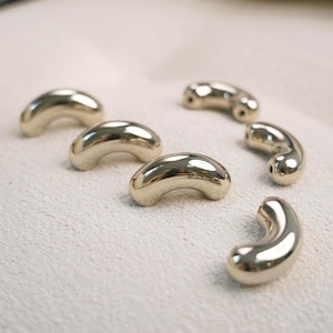 May include: A collection of six smooth, curved, metallic gold-toned beads. Each bead features a polished, reflective surface with a crescent shape and a small hole visible at the ends, designed for stringing in jewellery making projects.