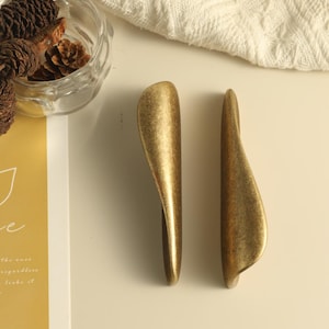 May include: Two elongated, curved cabinet pulls with a brushed, antique gold finish. The handles feature a smooth, wave-like silhouette with a textured metallic surface, positioned vertically side-by-side on a flat, light-coloured surface.