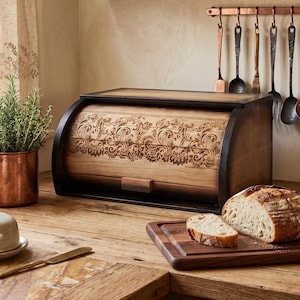 May include: A wooden bread box with a roll-top door featuring an intricate laser-engraved floral pattern. The box has dark brown side panels and a natural wood grain finish on the front, sitting on a wooden countertop next to a loaf of bread.