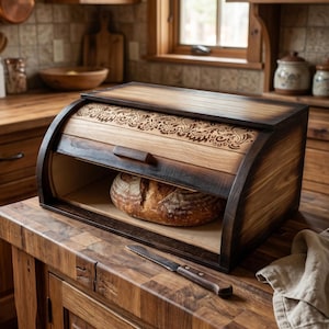 Op de afbeelding: Een houten broodtrommel met een roldeur voorzien van een gedetailleerd, lasergegraveerd bloemmotief. De trommel heeft een donker gebeitste afwerking en bevat een rond brood. Een metalen mes met houten handvat ligt op het aanrecht.
