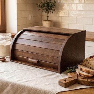 May include: A dark brown wooden bread box with a roll-top lid and a small horizontal handle. The rectangular container features horizontal slats across the curved front panel, providing a secure storage space for baked goods on a kitchen counter.