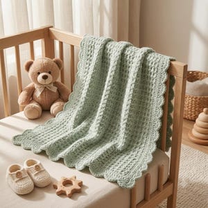 Könnte beinhalten: Eine hellsalbeigrüne gehäkelte Babydecke mit gewelltem Rand, ein brauner Plüschteddybär mit cremefarbener Schleife, ein Paar cremefarbene gehäkelte Babyschuhe und ein hölzerner Beißring in Sonnenform auf einer hellen Matratze.