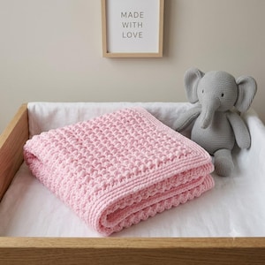 Pode incluir: Uma manta de bebé em croché cor-de-rosa claro com um padrão de textura de waffle e uma borda simples, acompanhada por um peluche de elefante em croché cinzento. A manta está dobrada cuidadosamente, exibindo o trabalho consistente.