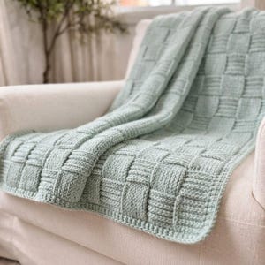 May include: A sage green knitted throw blanket featuring a prominent basketweave texture. The blanket is draped over a light-colored armchair, showcasing the thick yarn construction and the repeating square pattern of the woven knit design.