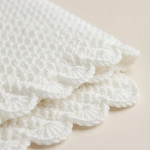 May include: A white crocheted textile featuring a dense, textured stitch pattern and a decorative scalloped edge. The fabric is folded, revealing multiple layers of the intricate, hand-worked yarn construction with a consistent, uniform finish.