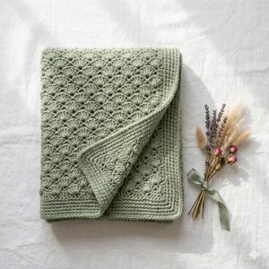 May include: A sage green crochet blanket featuring a repeating fan stitch pattern with a solid border. The blanket is folded to show the texture of the yarn work. A small bouquet of dried lavender and wheat stems tied with a ribbon is nearby.