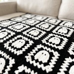 May include: A crochet blanket featuring a repeating pattern of square motifs. Each square consists of a white ghost figure centered within a black and white border. The textile has a textured, handmade appearance with consistent stitching throughout.