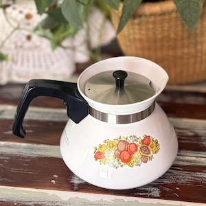 May include: A white teapot with a black handle and a silver metal lid. The body of the teapot features a colorful floral and fruit graphic print in shades of orange, yellow, and brown. The design includes a small black knob on the center of the lid.
