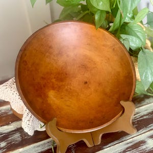 May include: A round, medium-brown wooden bowl with a smooth, polished interior surface and visible natural wood grain patterns. The bowl is displayed resting on a brown plastic stand, showcasing its circular shape and warm, consistent wood tone.