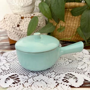 May include: A light turquoise ceramic saucepan with a matching lid and a single side handle. The piece features a smooth, glossy finish and a rounded body, designed for serving or heating small portions of food in a kitchen setting.