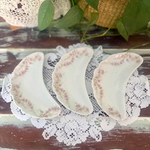 May include: Three white porcelain crescent-shaped bone dishes featuring a delicate scalloped edge and a floral pattern of small pink and yellow flowers along the curved rim. These individual serving dishes have a smooth, glossy finish.