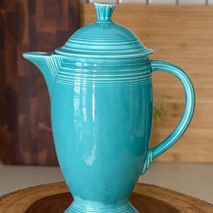May include: A turquoise ceramic coffee pot with a matching lid, featuring a tall, tapered body, a curved handle, and a pointed spout. The design includes horizontal ribbed detailing around the neck, the base, and the lid, with a glossy finish.