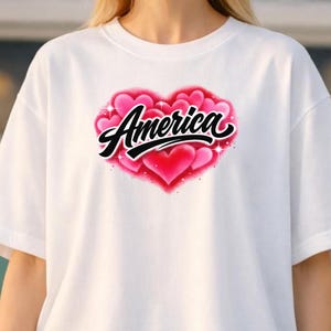 May include: A white short-sleeved t-shirt featuring a central graphic of a cluster of pink and red hearts airbrushed in a heart shape. The word America is printed in a bold black cursive script across the center of the heart design.
