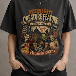 Pode incluir: Uma t-shirt cinza-carvão com uma estampa gráfica de quatro monstros clássicos do cinema comendo pipoca e hambúrgueres em um drive-in. O texto diz Midnight Creature Feature, Drive-In Theatre, Tonight Only, Admit One e Double Feature Tonight.