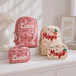 May include: Two corduroy backpacks and two matching rectangular pouches featuring a strawberry pattern. One set is pink with the name Luna in white script, and the other is cream with the name Maya in red script and small cherry appliques.