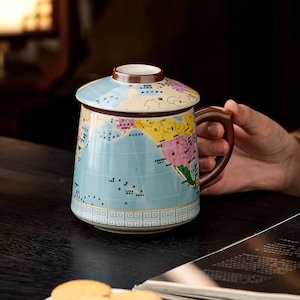 May include: A ceramic mug with a lid featuring a colorful world map print. The mug has a brown handle and a light blue base with a white geometric border pattern. The map displays various continents and oceans with text in Chinese characters.