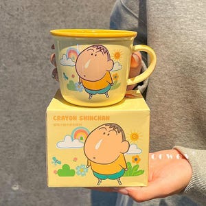 Könnte beinhalten: Ein gelber Keramikbecher mit einer Illustration der Zeichentrickfigur Crayon Shinchan, einem Regenbogen, einer Sonne und Blumen. Der Becher steht auf einer passenden gelben Pappschachtel mit dem Aufdruck Crayon Shinchan und chinesischen Schriftzeichen.