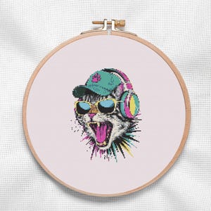Könnte beinhalten: Ein Kreuzstich-Stickprojekt in einem runden Holzrahmen mit einer verpixelten Katze, die eine türkisfarbene Baseballkappe mit rosa Pfotenabdruck, eine gelb umrandete Sonnenbrille und große rosa-türkisfarbene Kopfhörer trägt, mit buntem Spritzdesign.