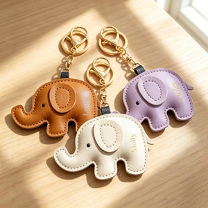 May include: Three elephant-shaped keychains made of smooth material, each featuring a gold-tone metal lobster clasp and ring. The keychains are brown, cream, and light purple. The cream one is embossed with the name Molly, and the purple one with Violet.