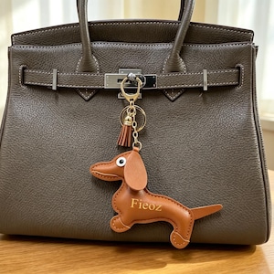 以下が含まれることがあります： シルバーの留め金とハンドルが付いた茶色の革製ハンドバッグ。 「Fieoz」という名前の茶色のダックスフンド型のキーホルダーがバッグに取り付けられています。 バッグは木製の表面に置かれています。