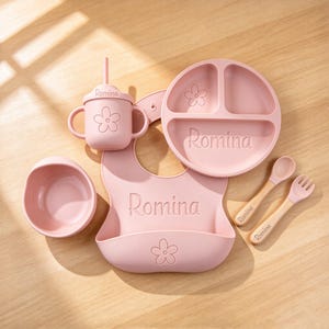 May include: A dusty pink silicone feeding set featuring a bib, a three-section plate, a bowl, a straw cup, a spoon, and a fork. Each item is embossed with the name Romina and a flower icon. The spoon and fork have light wood handles.