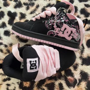 May include: A pair of black and light pink DC skate sneakers featuring thick light pink laces, a black suede upper with a white and pink floral graphic pattern, a pink DC logo on the side, and a silver cross charm attached to the eyelet area.