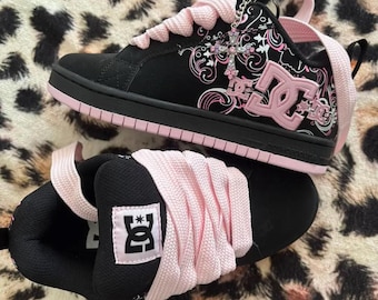 May include: A pair of black and light pink DC skate sneakers featuring thick light pink laces, a black suede upper with a white and pink floral graphic pattern, a pink DC logo on the side, and a silver cross charm attached to the eyelet area.