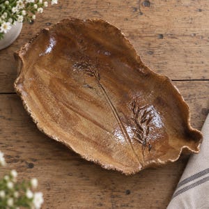 May include: A hand-formed ceramic tray shaped like a large leaf with a glossy, mottled brown glaze. The surface features a dark, pressed botanical imprint of a stalk of wheat or grass, with irregular, textured edges and a shallow basin.