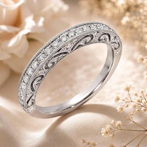 May include: A silver-toned metal band ring featuring a central row of small, round, clear stones set within a channel. The band is decorated with intricate, engraved scrollwork patterns along the sides and finished with a beaded milgrain edge.
