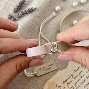 May include: A ring featuring a wide, smooth, pale pink band paired with a thin, silver-toned metal band set with a clear, oval-shaped gemstone. A small rectangular tag attached to the ring displays the text ARTISAN CRAFTED 75,000.