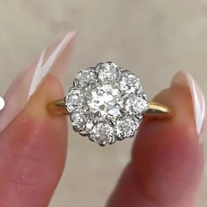 Op de afbeelding: Een gouden ring met een clusterzetting met een grote centrale ronde diamant omringd door acht kleinere ronde diamanten. De stenen zijn gezet in een zilverkleurige metalen kop, terwijl de ringband is vervaardigd uit geelgoud.