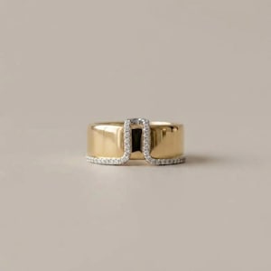 May include: A wide gold band ring featuring a U-shaped cutout detail on the top edge. The entire bottom edge and the inner perimeter of the U-shaped cutout are lined with small, clear, sparkling stones, creating a continuous shimmering border.