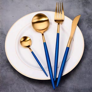 May include: A four-piece cutlery set comprising a small spoon, a large spoon, a fork, and a knife. Each item features a polished gold-tone metal head and a matte royal blue handle. The utensils are arranged on a white plate with a gold rim.