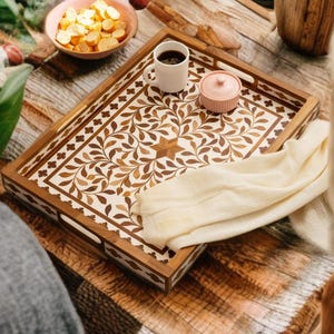 May include: A rectangular wooden serving tray featuring a white inlay with a brown leaf and vine pattern. The tray has integrated handles on the sides and holds a small white ceramic mug filled with black coffee and a small pink lidded container.
