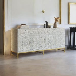 Puede incluir: Un aparador rectangular con un intrincado patrón floral incrustado en blanco y crema en sus paneles frontales y cajones. El mueble está sostenido por un marco de metal fino de tono dorado con cuatro patas e incluye pomos circulares transparentes.