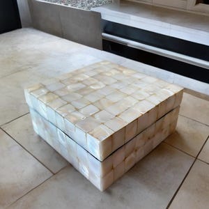 Könnte beinhalten: Eine quadratische Box mit Deckel, deren Außenfläche vollständig mit kleinen, schillernden, cremeweißen Perlmuttplättchen in einem Rastermuster bedeckt ist. Die Box hat eine flache Oberseite und gerade Seiten mit einer sichtbaren Naht.