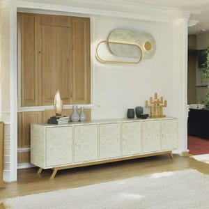 May include: A long, six-door sideboard cabinet featuring a light-colored, textured finish with subtle gold-toned patterns, supported by angled wooden legs. The cabinet surface holds several decorative objects, including vases and a gold-toned sculpture.