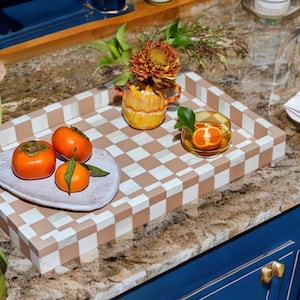 May include: A rectangular serving tray featuring a beige and white checkered pattern. On the tray sits a small yellow vase with a rust-colored flower, a light purple plate holding two bright orange persimmons, and a small glass bowl with a citrus slice.