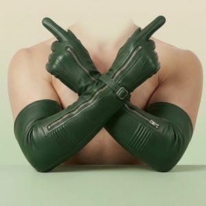 May include: A pair of long, dark green leather gloves extending past the elbows. The gloves feature a silver-toned metal zipper running along the top of the forearm, a buckled strap at the wrist, and ribbed detailing on the outer forearm area.