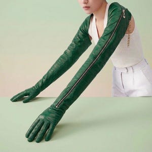 May include: A pair of dark green leather opera-length gloves featuring a full-length silver-toned zipper running along the outer arm. The gloves extend from the fingers up to the upper arm, showcasing a smooth texture and a fitted silhouette.