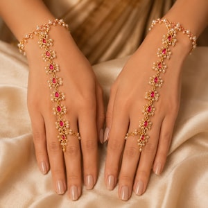 Könnte beinhalten: Ein Paar goldfarbene Handketten mit einem filigranen Design, verziert mit kleinen weißen Perlen und tropfenförmigen rosa Steinen. Jedes Schmuckstück verbindet ein Armband mit einem Ring und bildet eine dekorative Kette über dem Handrücken.