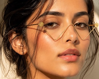 May include: Gold-toned wire glasses featuring a distinctive V-shaped bridge and thin metal frames. The temples are accented with small, circular flower-shaped details at the hinges, creating a delicate and structural look for these eyewear frames.