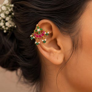 May include: A gold-toned ear cuff featuring a crescent-shaped design with vibrant pink and green stones. The piece is accented with small white pearls and dangling green beads, creating a decorative ornament that sits along the ear cartilage.
