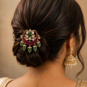 May include: A gold-toned hair accessory featuring a crescent moon shape with a central green stone, surrounded by clear crystals and a deep pink band. Five dangling green oval beads hang from the bottom. A gold bell-shaped earring with small white pearls hangs from the ear.