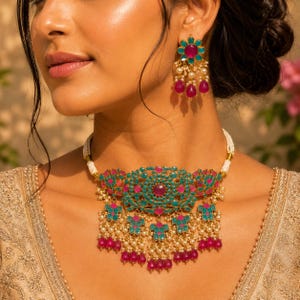Könnte beinhalten: Ein goldfarbenes Choker-Halsband und passende Ohrhänger mit türkisfarbenen und magentafarbenen Steinen. Die Halskette hat einen breiten, blumenartigen Mittelanhänger mit baumelnden magentafarbenen Perlen und kleinen weißen Perlen an einer Kordel.